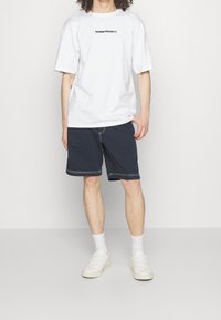 Camiseta de manga corta blanca con el texto "DENIM PROJECT", combinada con pantalones cortos azul marino con costuras blancas y zapatillas blancas.