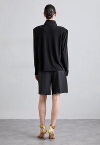 Black long-sleeve shirt with a collar, paired with black shorts and gold high-heeled sandals. Smooth materials, minimalist design.