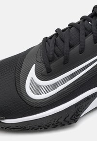 Nike-sko med en sort syntetisk overdel med en struktureret finish, hvid swoosh-logo og en mønstret gummisål. Der er detaljer som "PRECISION 7."