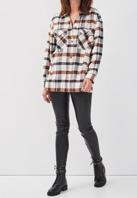 Chemise à carreaux en crème, marron et marine ; tissu doux, fermeture à boutons sur le devant, deux poches poitrine. Associée à des leggings en cuir noir et des bottes.