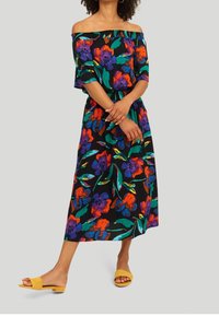 Robe noire décolletée avec un imprimé floral coloré en rouge, violet et vert ; taille élastique ; associée à des sandales jaunes à enfiler.