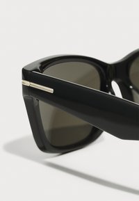 Gafas de sol de plástico negro con lentes oscuros, forma angular y un detalle dorado en la patilla. Acabado brillante en la montura.