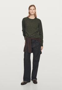 Olivengrøn sweater med lange ærmer, brun strikket sweater bundet om taljen, kombineret med mørkeblå, vidt ben jeans og brune loafers.