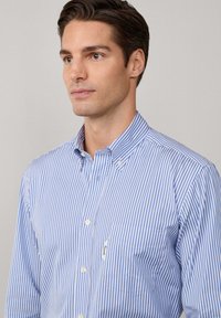 Joven vestido con una camisa de botones a rayas verticales en azul y blanco, con un bolsillo en el pecho izquierdo, mirando ligeramente hacia un lado.