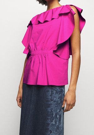 Frau trägt ein leuchtend pinkes, ärmelloses Rüschen-Top mit geraffter Taille, kombiniert mit einem dunkelblauen Jeansrock mit Blumenmuster.