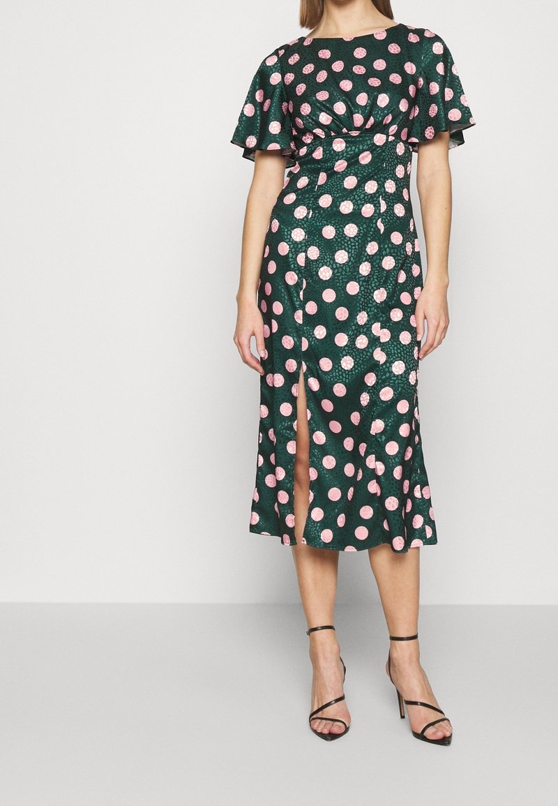Grön midi-klänning med prickar, volangärmar, rynkat liv, sidslits och mjuk textur. Bärs med svarta sandaletter med remmar.