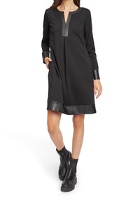 Femme portant une robe noire à manches longues avec des garnitures en cuir au niveau du col, des poignets et de l'ourlet, ainsi que des bottines noires, debout avec une main dans la poche.