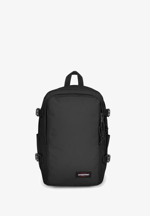 Mochila de tela negra con forma rectangular, correas ajustables, bolsillos laterales y una etiqueta distintiva en la parte frontal. Superficie texturizada.