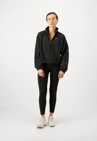 Pullover in pile nero con collo con zip, vita elastica e polsini; abbinato a leggings neri e scarpe da ginnastica di colore chiaro.