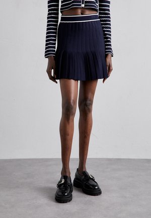 Osoba ubrana w granatową plisowaną spódnicę, czarne mokasyny z klamrą oraz krótki czarno-biały sweter w paski z długimi rękawami.