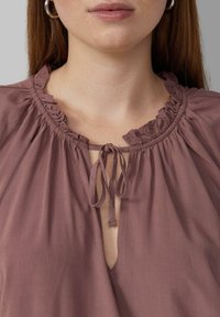 Pudrowa bluzka w kolorze mauve z falbaną przy dekolcie, z przodu z zapięciem na sznurek i luźnym krojem. Miękka tkanina o gładkiej fakturze.