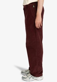 Burgundy corduroy pants with a straight shape, featuring a green label on the waistband, paired with grey athletic shoes.