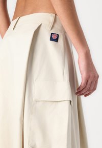 Beige pleated skirt featuring a large side pocket and a "Relax" logo patch at the back waistband. Smooth, lightweight fabric texture.