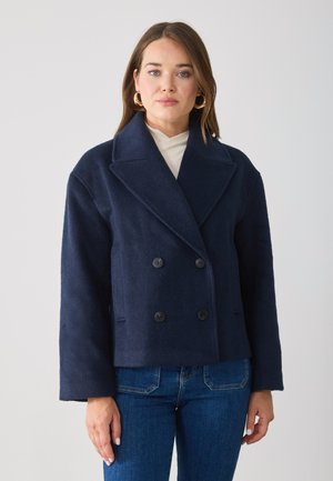 Giacca doppiopetto in misto lana blu navy con grandi rever, due tasche frontali e bottoni neri, abbinata a una maglia crema e jeans blu.