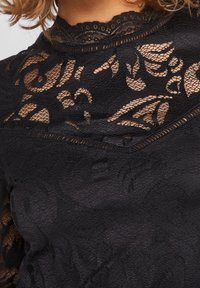 Sort blonde top med blomster mønstre, tekstureret bund, høj hals og intrikate bølgede kanter. Elegante design med gennemsigtige elementer.