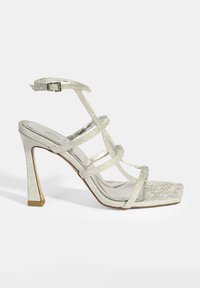 Glatte High-Heel-Sandale mit Riemchen in silbernem Glitzer und quadratischer Spitze. Verfügt über eine glänzende Oberfläche, einen verstellbaren Knöchelriemen und ein Blockabsatzdesign.