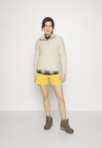 Beige fleece trøje med halv lynlås, gule shorts og brune vandresko. Fleecen er blød, og trøjen har en brystlomme.