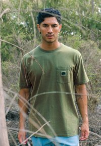 Olive green cotton t-shirt with a chest pocket featuring an elephant logo. Relaxed fit and short sleeves, paired with light blue jeans.