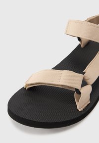 Beige stoffrem sandaletter med en teksturert svart gummisåle, med justerbare remmer og en enkel, minimalistisk design.