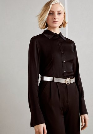 Blonde vrouw die een zwart jumpsuit met knopen en lange mouwen draagt, met een zilveren riem en creolen, staand tegen een effen achtergrond.