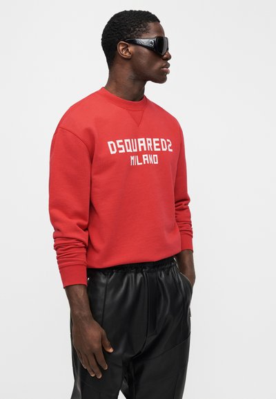 Hombre con gafas de sol negras, sudadera roja con el texto "DSQUARED2 MILANO" y pantalones de cuero negros con las manos en los bolsillos.