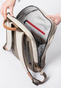 Zaino beige e marrone con dettagli in pelle, dotato di un interno grigio con una tasca con zip rossa e slot organizzativi per accessori.
