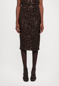 Mujer que lleva una falda de lentejuelas marr�ón oscuro hasta la rodilla, medias negras transparentes y sandalias de tacón alto negras con tiras, todo sobre un fondo blanco.