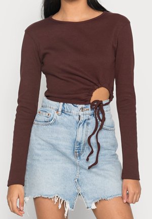 Femme portant un haut marron à manches longues avec une découpe froncée et un lien sur le côté, assorti à une jupe en jean bleu clair à taille haute et bords effilochés.