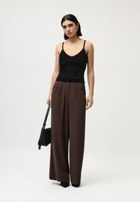Čierny textúrovaný top s ramienkami a detailom mašle, skombinovaný s vysokým pásom a širokými hnedými nohavicami. Modelka drží čiernu crossbody tašku.
