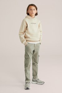 Dítě stojící s rukama v kapsách, oblečené v béžové mikině s kapucí, zelených cargo kalhotách a šedých teniskách New Balance na neutrálním pozadí.