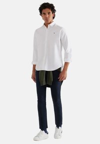Camicia bianca con maniche arrotolate, pantaloni blu navy, maglione verde annodato in vita e sneakers bianche con dettagli blu navy.
