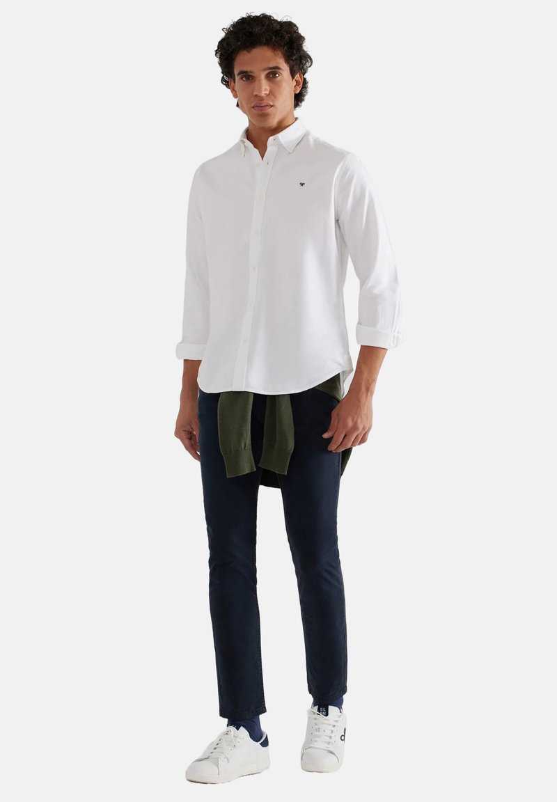 Camicia bianca con maniche arrotolate, pantaloni blu navy, maglione verde annodato in vita e sneakers bianche con dettagli blu navy.