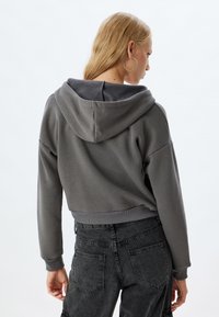 Sudadera gris corta con capucha, que presenta una textura suave y puños acanalados. Combinada con jeans oscuros de cintura alta, exhibiendo un estilo casual.