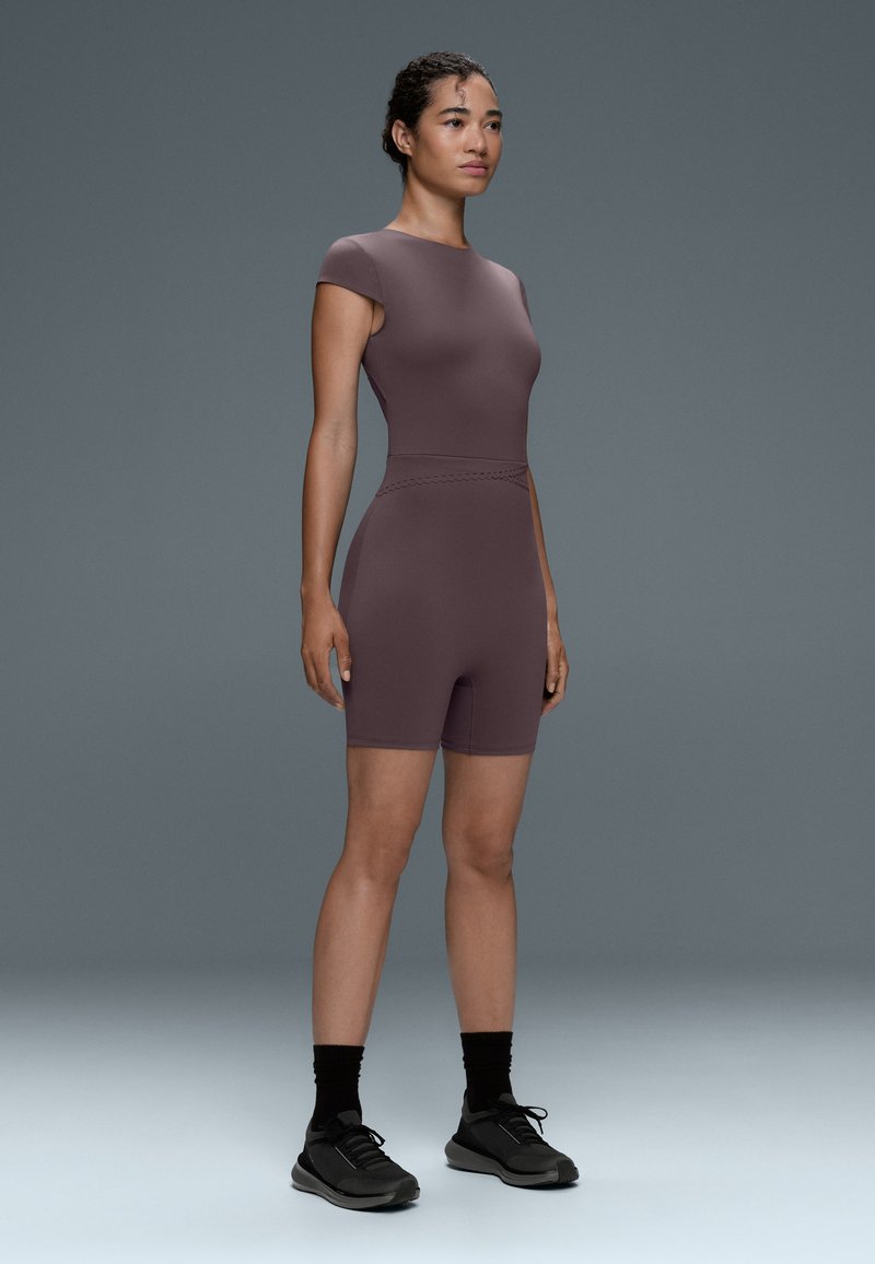 Femme debout, portant une combinaison courte mauve ajustée, des chaussettes noires et des chaussures de sport noires sur un fond gris uni.