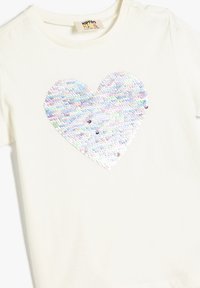 Hellbeige Baumwoll-T-Shirt mit großem, pastellfarbenem Paillettenherz-Design in der Mitte. Runde Ausschnitt, kurze Ärmel.