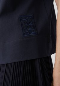 Top in cotone navy con un inserto ricamato a trame con motivo a foglia sul lato sinistro, abbinato a una gonna scura plissettata.