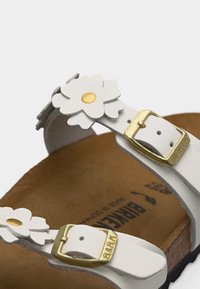 Λευκές λουρίδες σανδαλιών με χρυσά αγκράφες και λευκές διακοσμήσεις λουλουδιών σε ταμπέ κωκ πόδι με ορατό λογότυπο.