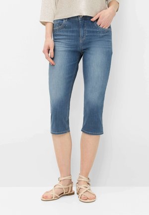 Kvinde iført lyseblå cropped denim jeans med nittebesatte forlommer og beige flade sandaler med stropper, stående mod en hvid baggrund.