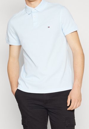 Mann trägt ein hellblaues Polohemd mit kurzen Ärmeln und kleinem Logo auf der Brust sowie schwarze Cargohosen, eine Hand in der Tasche.
