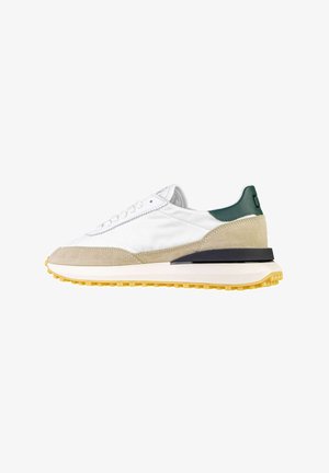 Sneaker bianca con dettagli in suede beige, linguetta verde sul tallone, particolare della suola intermedia nera e suola in gomma gialla, mostrata di profilo su sfondo bianco.