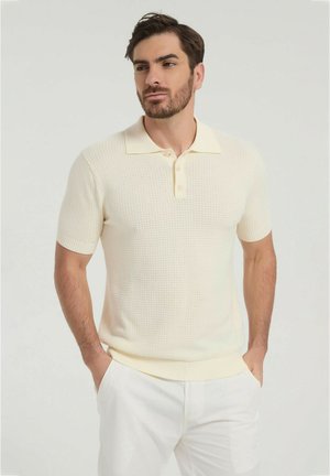 Homme barbu portant un polo crème texturé à manches courtes et un pantalon blanc, debout les mains dans les poches devant un fond uni.
