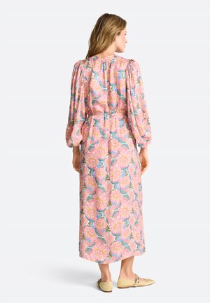 Frau, die rückwärts blickt und ein rosa, langärmeliges Midikleid mit Blumenmuster, Puffärmeln und beige flache Schuhe trägt.