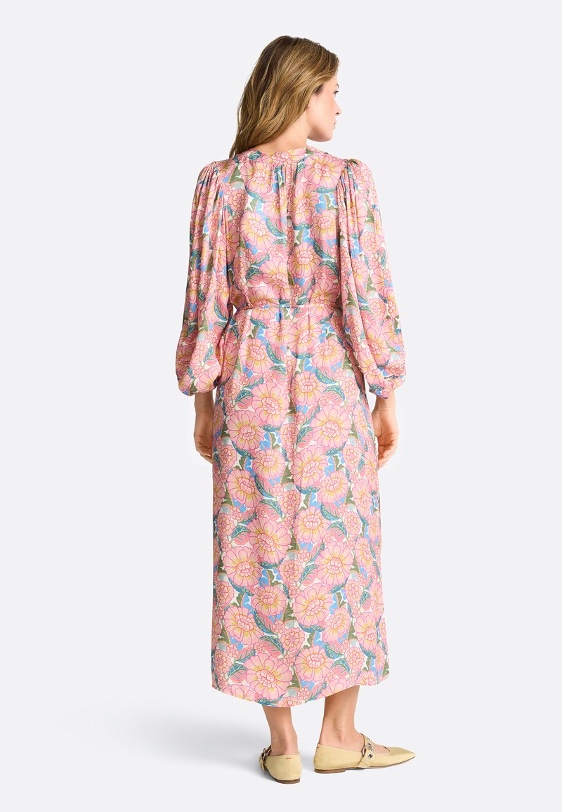 Femme vue de dos portant une robe midi à manches longues à fleurs roses avec des manches bouffantes et des chaussures plates beiges.