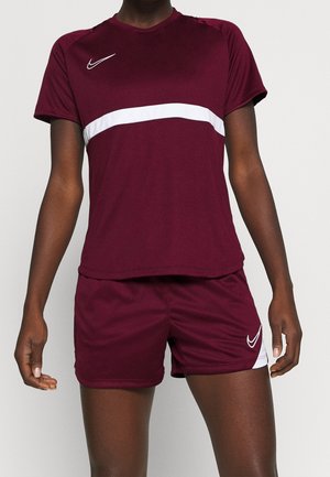 Osoba v bordovém sportovním oblečení s bílým vodorovným pruhem na tričku a bílými logy Nike na tričku i šortkách.