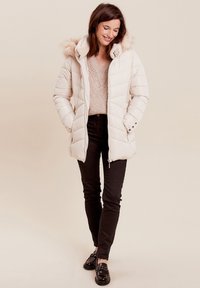 Helle beige Daunenjacke mit einer mit Fell gefütterten Kapuze, gestepptem Design, Reißverschluss und Druckknopfverschlüssen, kombiniert mit einem gestrickten cremefarbenen Pullover und schwarzen Hosen.