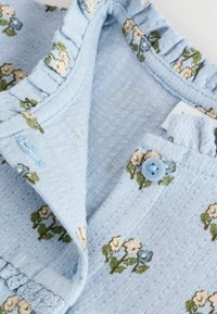 Blå blommig klädsel med ribbad textur, volangkrage och en ljusblå knapp. Broderade blommor i rosa, grönt och blått.