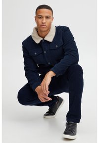 Chaqueta de pana azul marino con un cuello de piel sintética color crema, dos bolsillos frontales, combinada con pantalones azul marino oscuro y zapatillas negras con cordones.