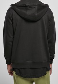 Sudadera negra con capucha, con puños y dobladillo acanalados, que presenta un diseño en capas y una tela texturizada. La vista trasera muestra un acabado suave.