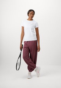 Kratka bela majica s malim logotipom, v kombinaciji z ohlapnimi burgundskimi trenirkami. Tenisice so bele s roza poudarki. Drži tenis raketo.
