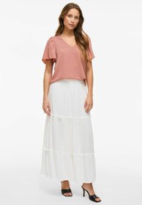 Blusa rosa pallido con maniche corte a campana e scollo a V, abbinata a una gonna maxi bianca a strati. Sandali neri con tacco alto completano l'outfit.